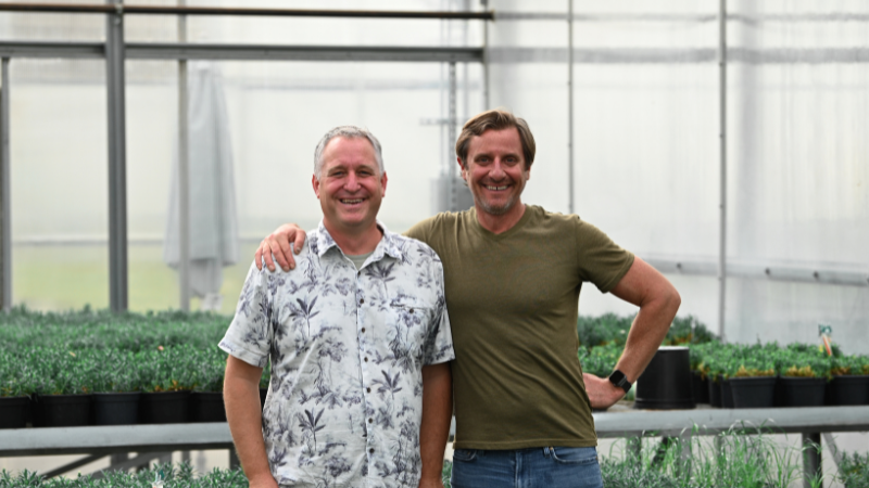Two men standing in a greenhouse with one arm around the others should for the photo