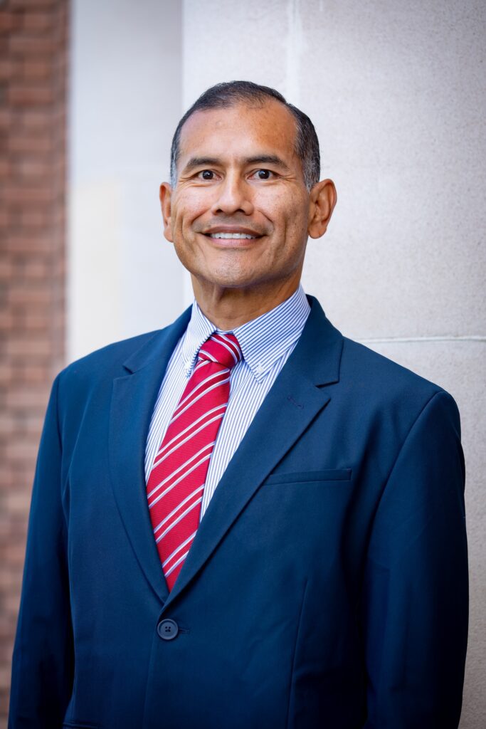 Headshot of man smiling and wearing a blue suit jacket, light blue shirt and red tie with stripes
