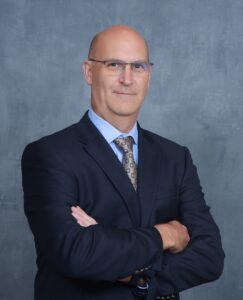 Head shot of man in dark blue suit with arms crossed, wearing glasses